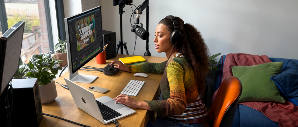 Seorang perempuan sedang bekerja di rumah di mejanya dengan MacBook Pro yang terhubung ke dua layar eksternal, mengenakan headphone, dan berbicara melalui mikrofon