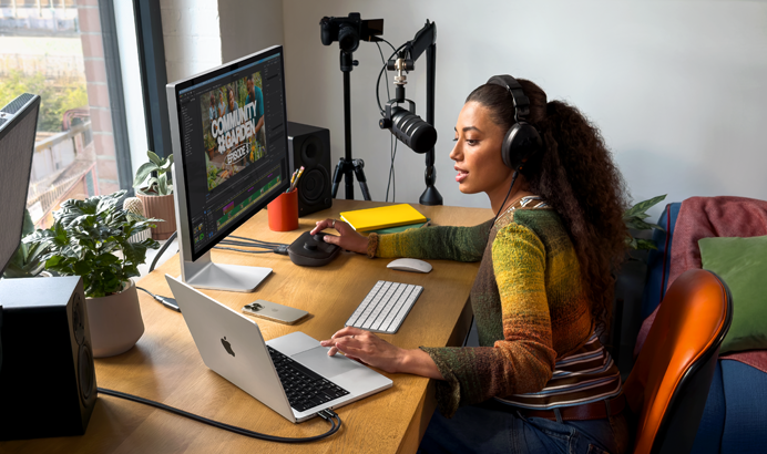 Seorang perempuan sedang bekerja di rumah di mejanya dengan MacBook Pro yang terhubung ke dua layar eksternal, mengenakan headphone, dan berbicara melalui mikrofon