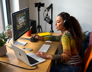 Seorang perempuan sedang bekerja di rumah di mejanya dengan MacBook Pro yang terhubung ke dua layar eksternal, mengenakan headphone, dan berbicara melalui mikrofon